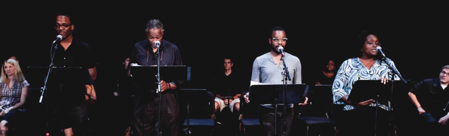 Four performers standing in front of microphones and music stands with an audience seated behind them against a black background