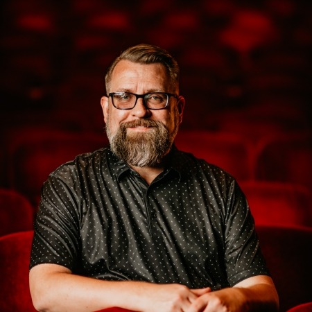 man with brown hair with beard and glasses, grey shirt sitting in red theater seats