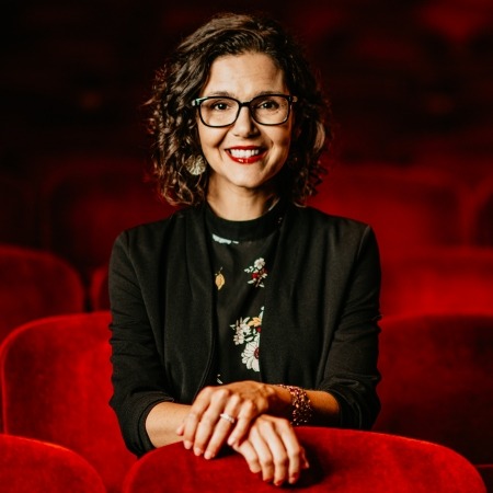 Woman with brown hair, glasses, black blazer sitting in red theater seats