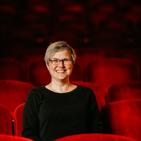woman with grey hair, glasses and black blouse sitting in red theater seats