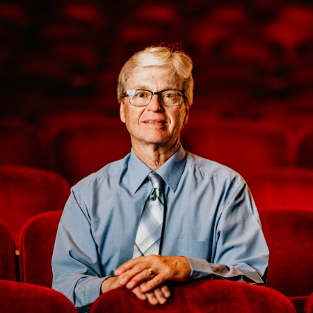 Man with white hair, black glasses, blue button up and tie sitting in red theater seats
