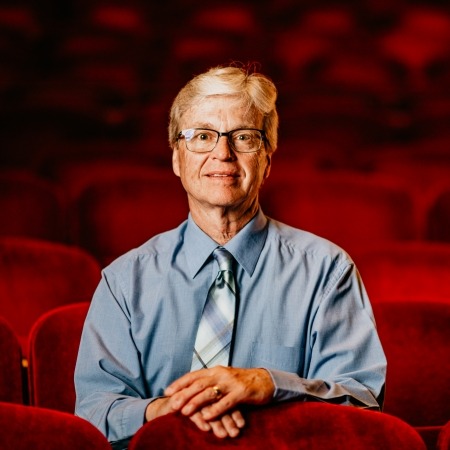 Man with white hair, black glasses, blue button up and tie sitting in red theater seats