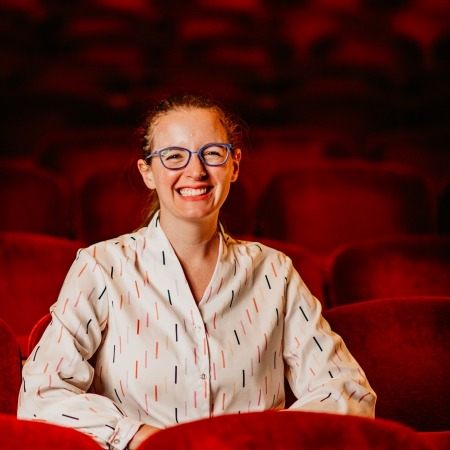 woman with brown hair, blue glasses, multi colored blouse sitting in red theater seats