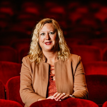 Woman with loose curls blonde hair in a tan leather jacket sitting in red theater seats