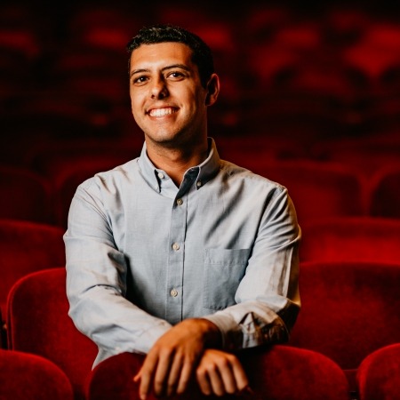 Man in blue button up with black hair sitting in red theater seats