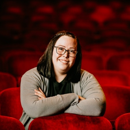 women with black hair, black glasses, grey cardigan, black blose sitting in red theater seats