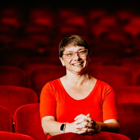 Woman with brown hair, glasses, red blouse sitting in red theater seats