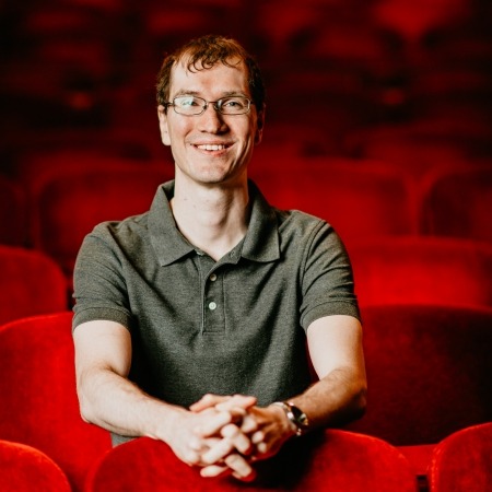 man with brown hair, gray shirt, glasses sitting in red theater seats