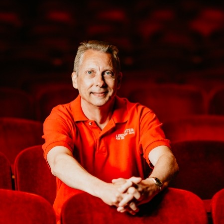 man with red shirt and grey hair sitting in red theater seats