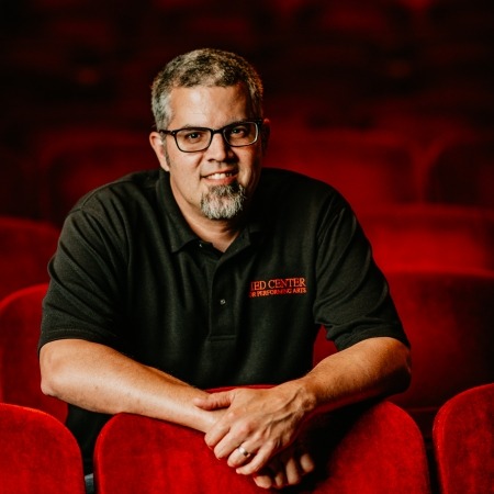 man with brown grey hair with black glasses and a beard in a black shirt sitting in red theater seats