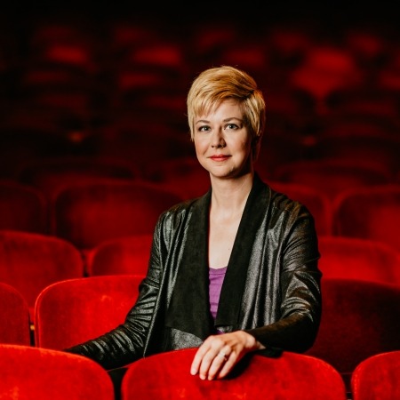 woman with dirty blonde hair with a black blazer and purple blouse sitting in red theater seats