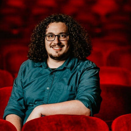 man with black curly hair in teal button up sitting in red theater seats