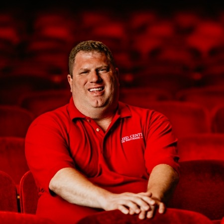 man with brown hair and red shirt sitting in red theater seats