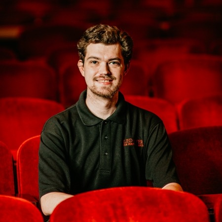 man with brown hair and black shirt sitting in red theater seats