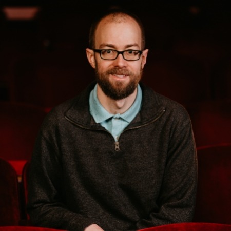 man with brown hair with black glasses and brown beats in sky blue shirt underneath a quarter zip sitting in red theater seats