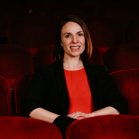 Woman with brown  hair in a black blazer and red blouse sitting in red theater seats