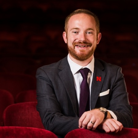 man with ginger hair with mustache and beard, in a suit  sitting in red theater seats